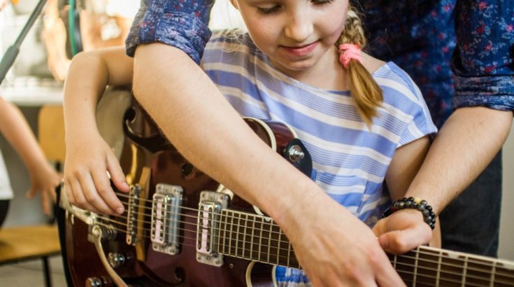 Uśmiechnięta dziewczynka w koszulce w niebieskie paski trzyma w ręku gitarę elektryczną. Nad nią widoczna ręka instruktora, który pomaga jej ustawić palce na strunach.