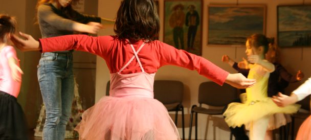 Zajęcia baletowe. Na pierwszym planie dziewczynka w różowej spódniczce, z wyciągniętymi w bok rękami, widoczna od tyłu. W tle inne dzieci i instruktorka zajęć w baletowej pozie.