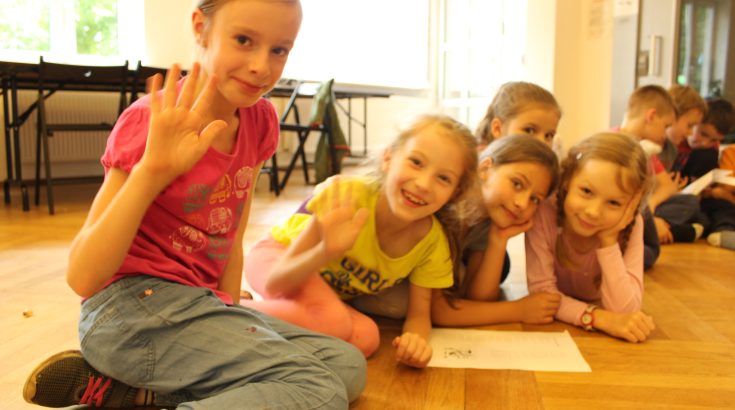 Grupa pięciu dziewczynek siedzi na podłodze Klubu Kultury. Przed nimi kartka papieru. Dziewczynki uśmiechają się i machają do obiektywu.