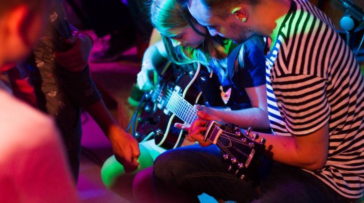 Młoda dziewczyna trzyma gitarę elektryczną. Po prawej stronie instruktor. Wokół kolorowe światła.