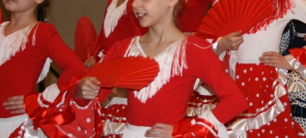 Zbliżenie na grupę dziewczynek w czerwono-białych strojach do flamenco, z czerwonymi wachlarzami.