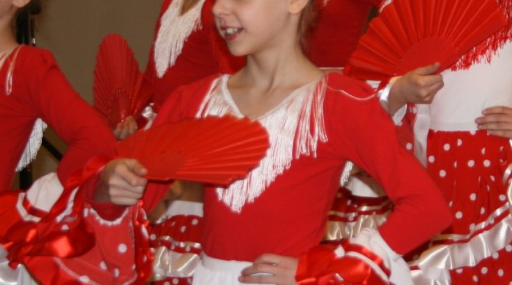 Zbliżenie na grupę dziewczynek w czerwono-białych strojach do flamenco, z czerwonymi wachlarzami.
