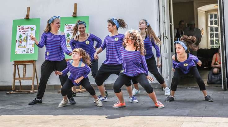 Zespół siedmiu młodych dziewczyn w fioletowych koszulkach wykonuje układ taneczny.