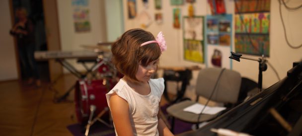 Ciemnowłosa dziewczynka w białej bluzce i różowej opasce na głowie siedzi przy fortepianie. Za nią widoczne inne instrumenty.