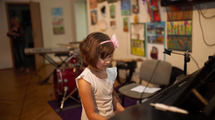 Ciemnowłosa dziewczynka w białej bluzce i różowej opasce na głowie siedzi przy fortepianie. Za nią widoczne inne instrumenty.