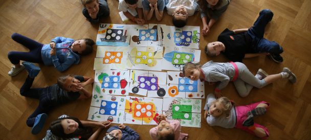 Widoczna z góry grupa dzieci siedzi na podłodze wokół kartek z kolorowymi rysunkami.