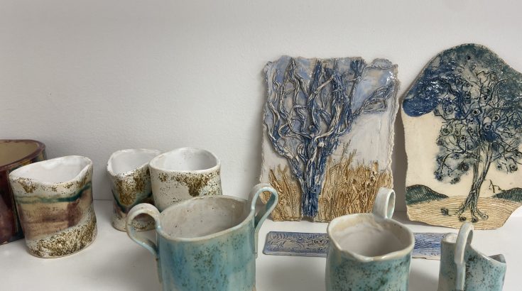 6 kubków ceramicznych beżowo, - zielono- niebieskich oraz dwie płaskorzeźby przedstawiające drzewa również w kolorze beżowym, zielonym i niebieskim. Wszystko ustawione jest na białym tle.